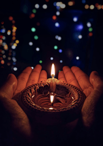 Hands gently cradle a lit clay lamp against a dark background, with colorful, blurred lights in the distance, evoking warmth and celebration.