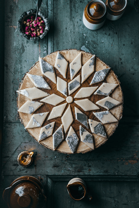 A wooden table holds a round tray of intricately arranged diamond-shaped sweets, some with silver foil. Nearby are jars, a small dish, and cups, creating a warm, inviting scene.