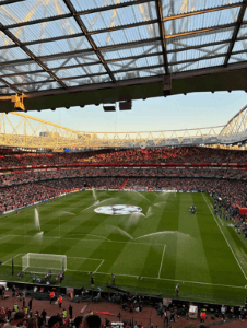 A large stadium filled with cheering fans. A football pitch displays the UEFA logo at the center, with sprinklers watering the grass.