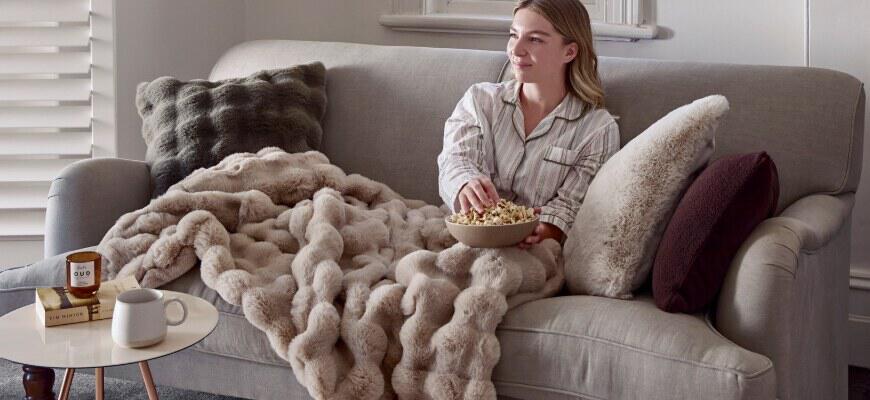 Bed Bath N Table deals page image showing a woman suggled up on a sofa with a warming blanket