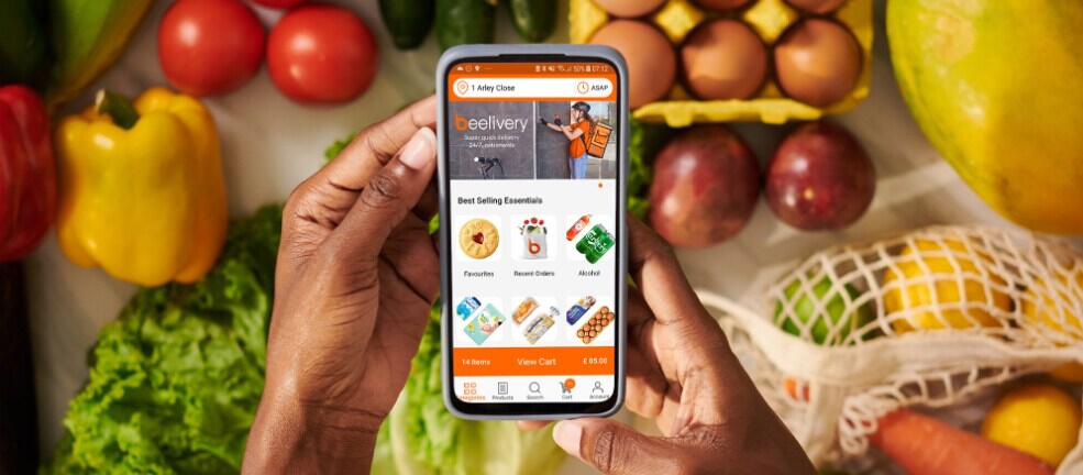Beelivery savings page image - Hands holding a smartphone shopping the Beelivery app with groceries, including vegtables in the background