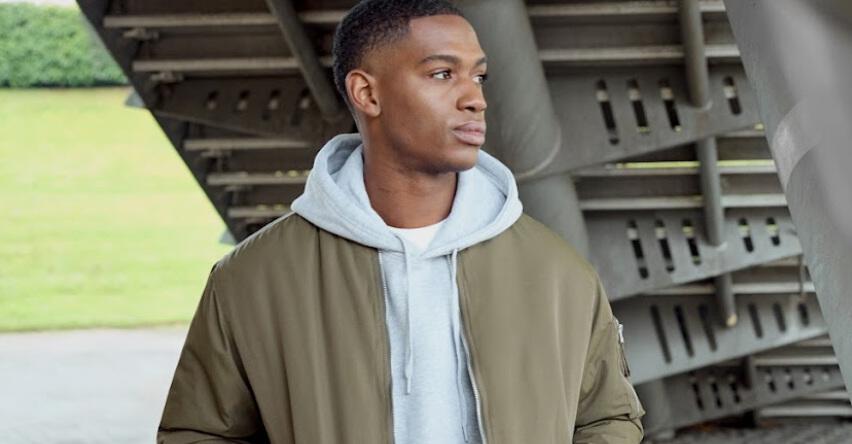 Burton new season menswear image - male model wearing a grey hoodie and green jacket combo in an idustrial setting