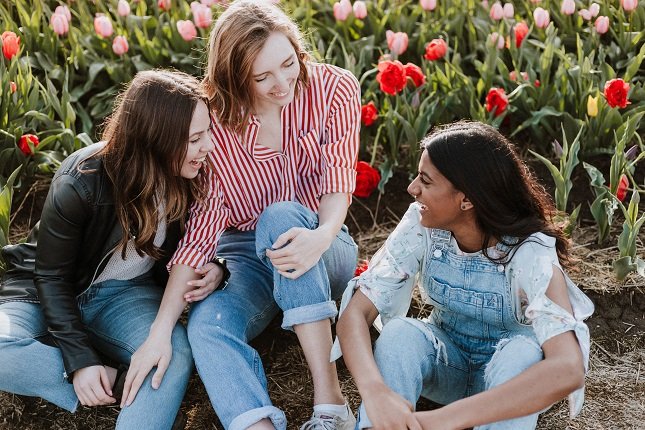 Freundesgruppe von drei Studentinnen sitzt lachend in einer Blumenwiese