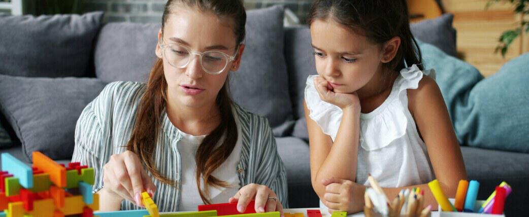 A woman sitting next to a young girl while showing her how to assemble a construction toy