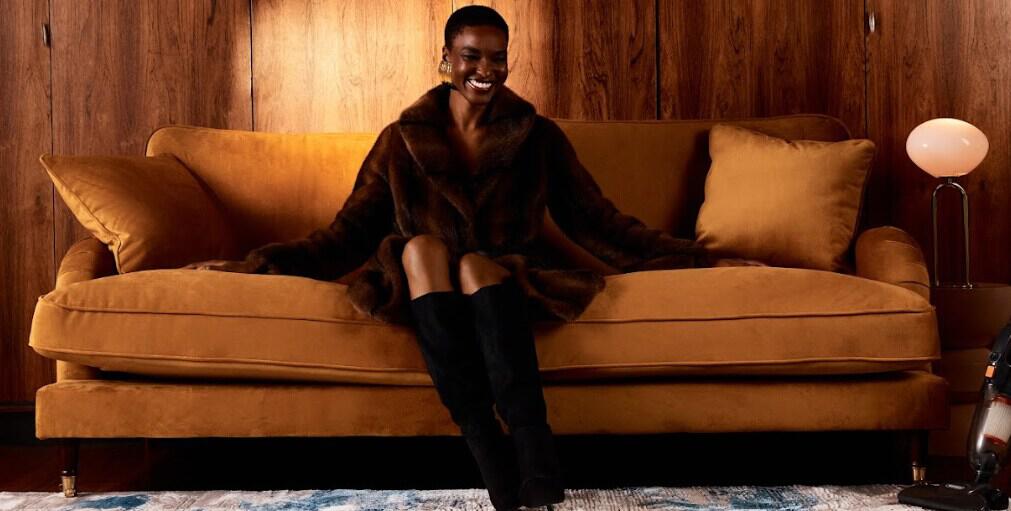 Debenhams new season image showing a woman in brown coat and black boots sitting on a sofa