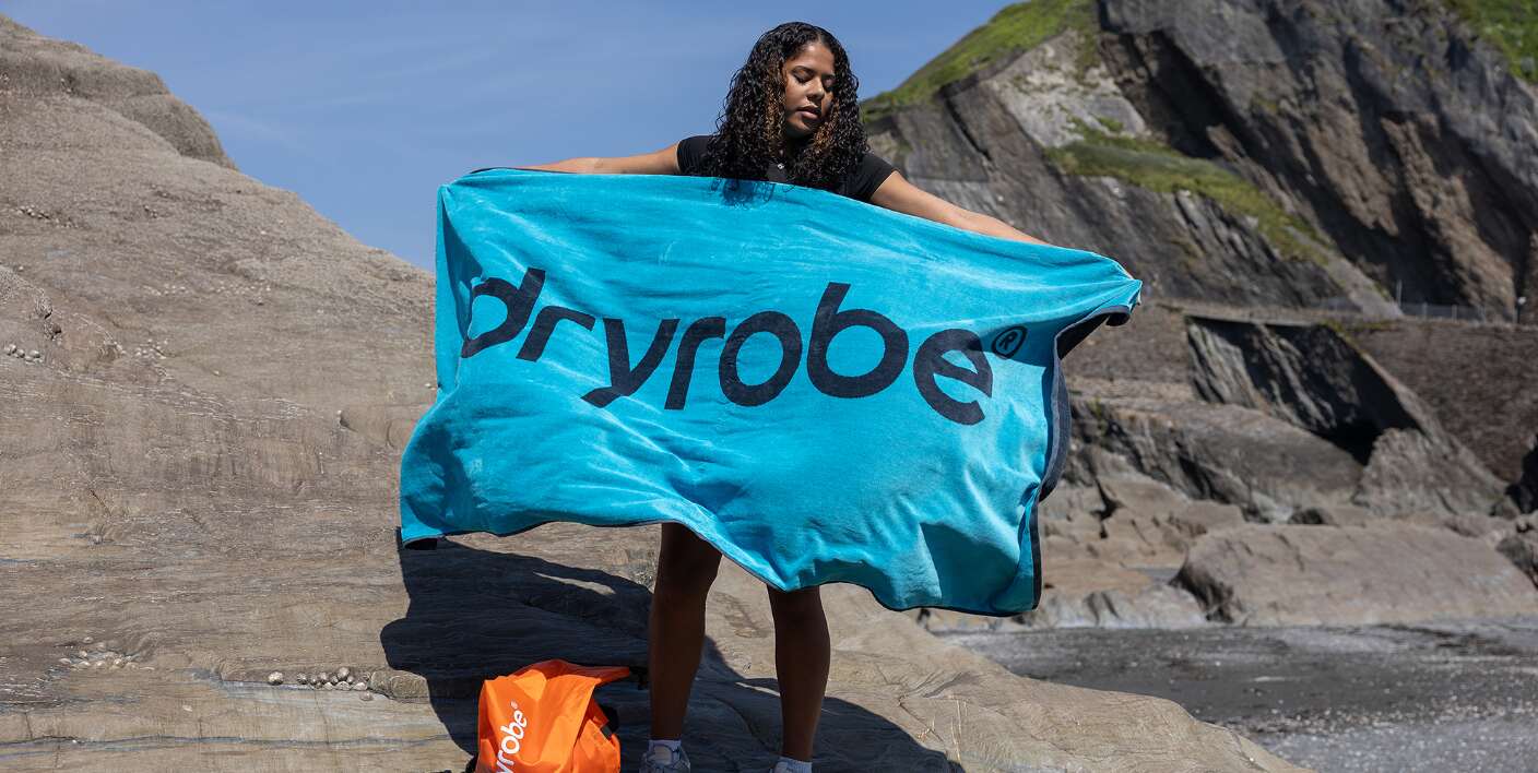 Dryrobe offers page banner - A woman on a beach holding out a blue towel with Dryrobe printed in big letters on it