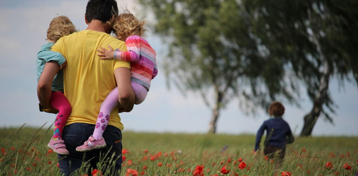 family holiday picture showing a dad carrying two children through a field,while another one runs ahead