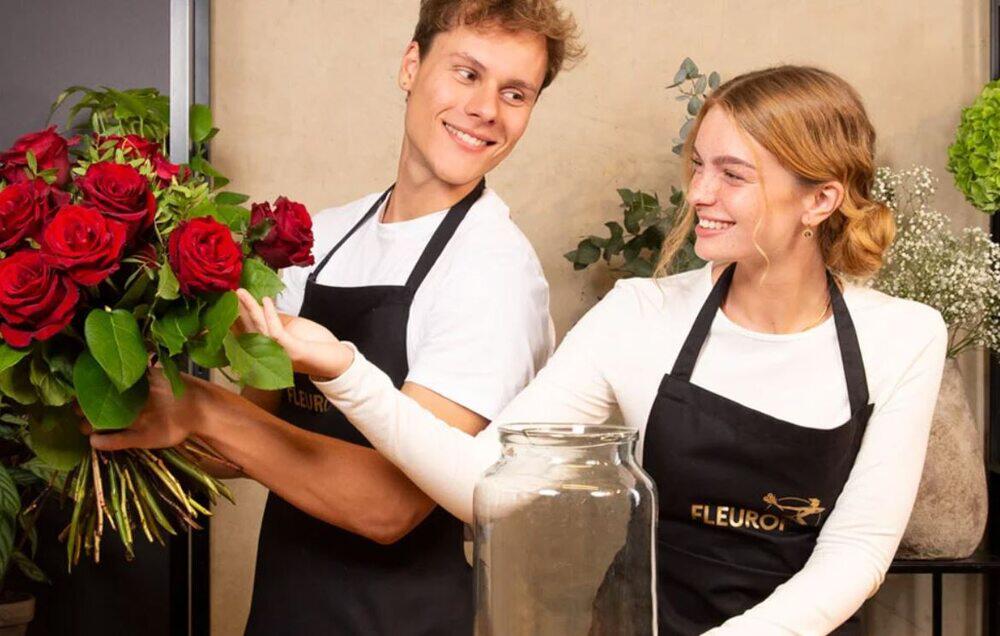 Junger Florist hält einen Fleurop Rosenstrauß, junge Floristin in Fleurop Schürze fasst den Rosenstrauß sanft an