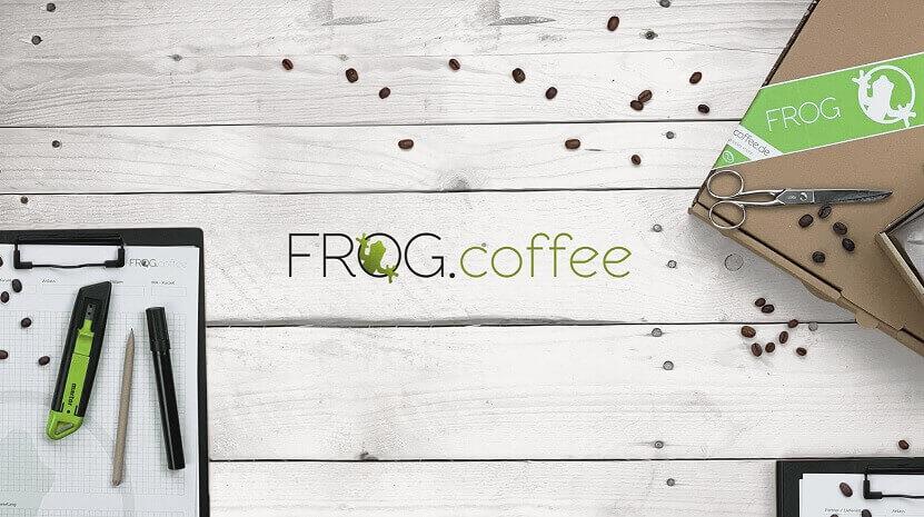 Firmenlogo mir Kaffeebohnen, einem Versandkarton sowie Schreibutensilien auf einem Tisch als Banner für die FROG.coffee Gutscheinseite.