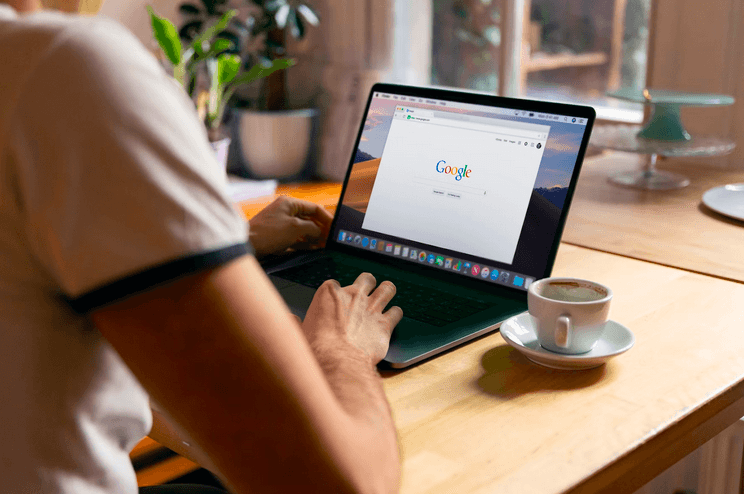 Mann sitzt in einem Café und hat seinen Laptop mit der Google Startseite vor sich um aktuelle Finanzfragen zu recherchieren