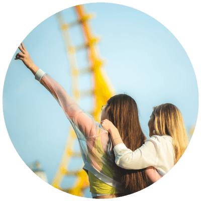 two girls at a theme park. One girl has her hand in the air, with rollercoaster track in the background