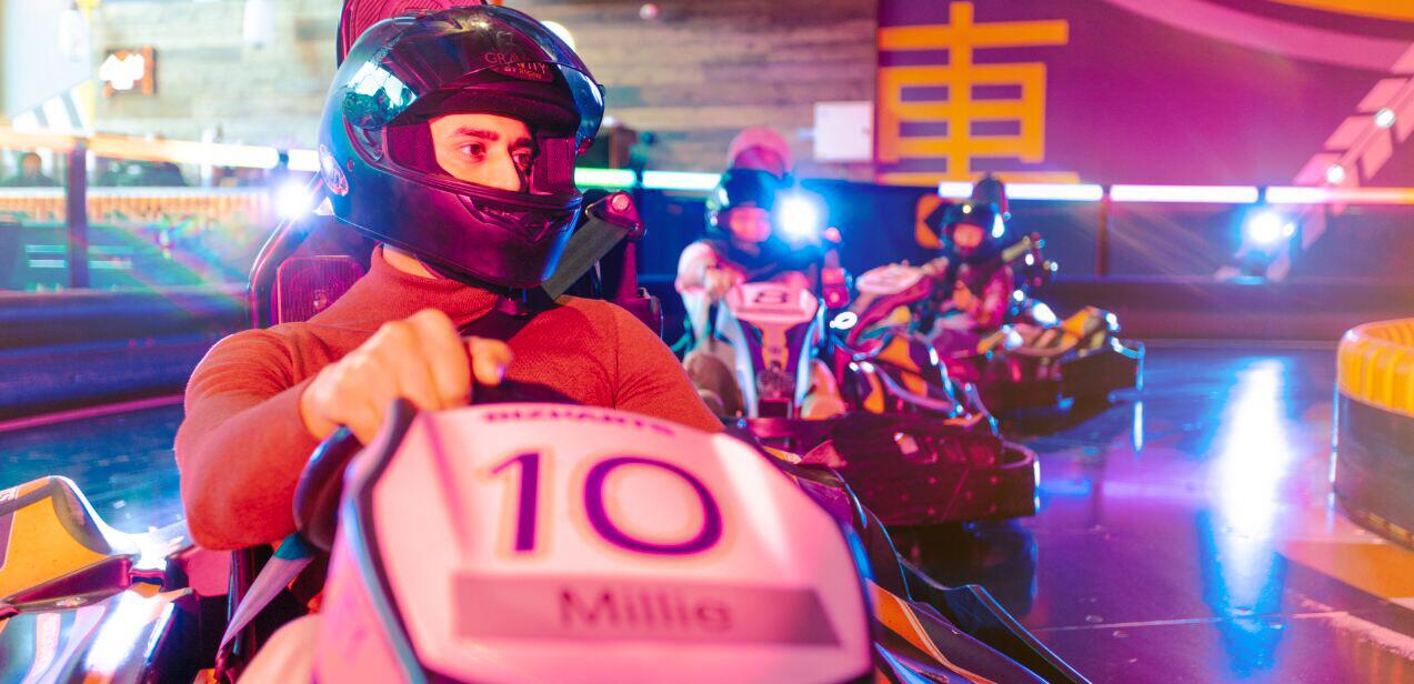 Gravity how to save money guide image - A close up shot of a man go-karting, with others racing in the background