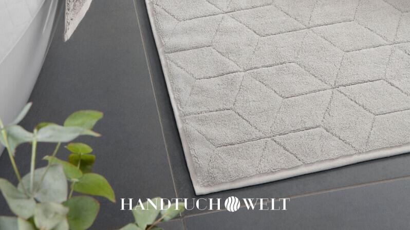 Banner der Handtuch Welt Gutscheinseite: eine graue Badezimmermatte auf dunkelgrauen Fliesen neben einer Wanne und einer Pflanze