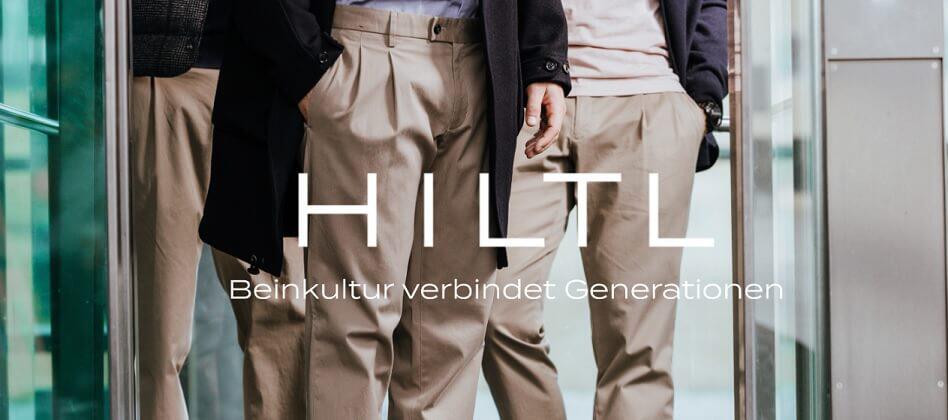 Banner der Hiltl Gutscheinseite: drei Herren in beigefarbenen Herrenhosen stehen in einer geöffneten Glastür; Bildausschnitt zeigt nur die Beine und einen Teil des Oberkörpers; Aufschrift "Hiltl – Beinkultur verbindet Generationen"