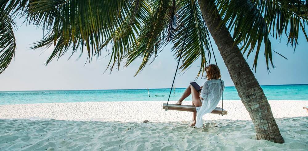 Banner für die HLX Gutscheinseite: Frau mit Buch auf einer Schaukel, die am Strand von einer Palme herabbaumelt