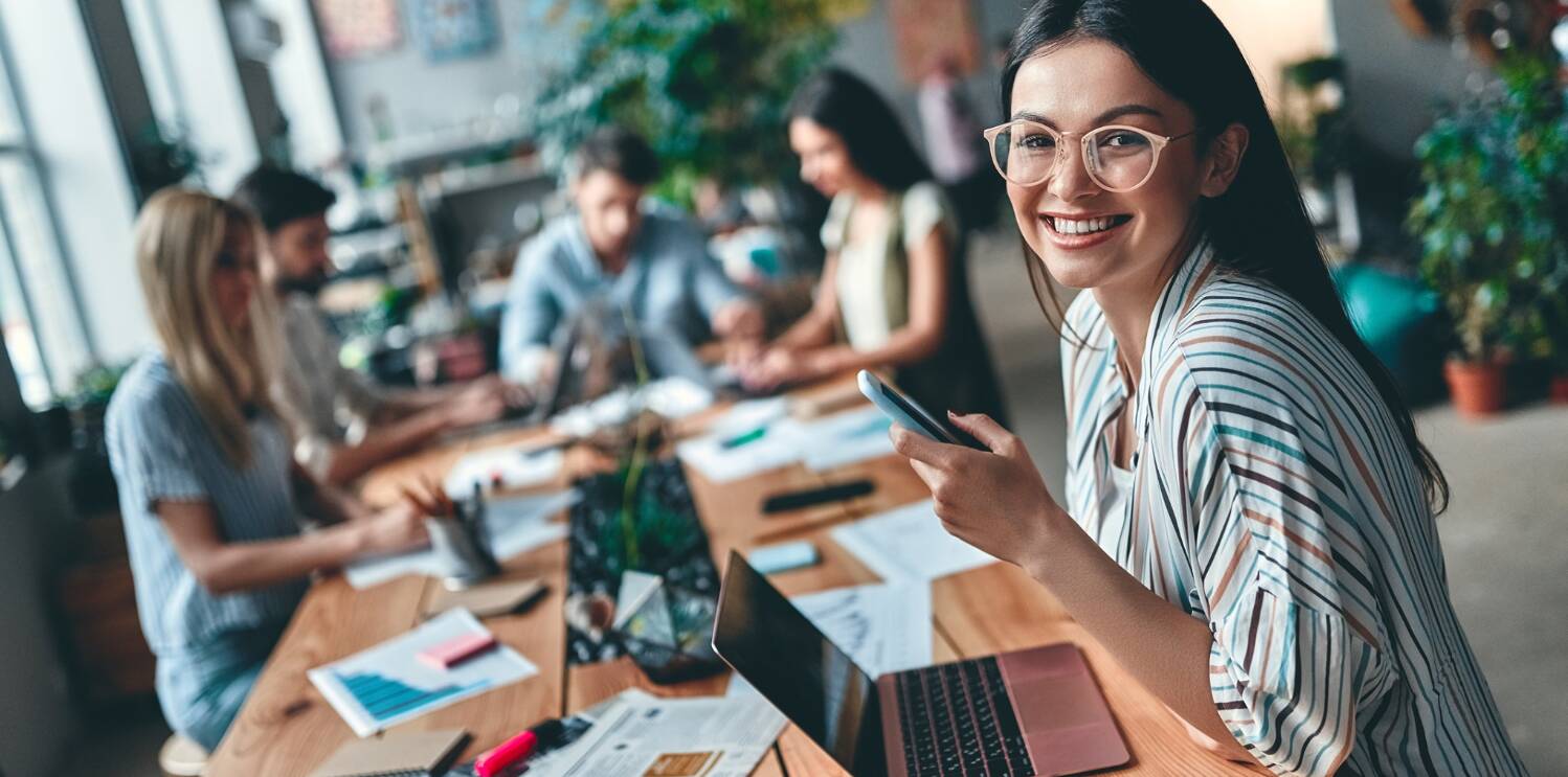 International Open Academy savings banner - Image showing a woman smiling while holiday a smartphone and using a laptop. Other people wording at a desk can be seen in the background