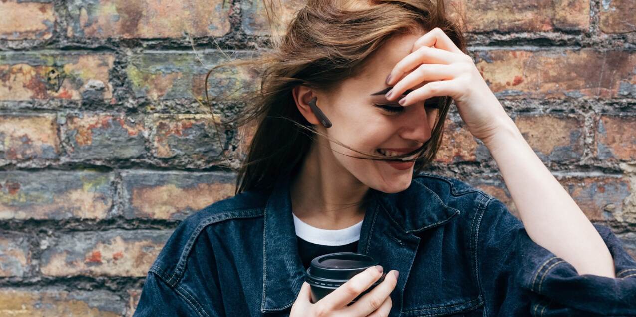 JBL deals page banner - A woman wearing earbuds and laughing, with brick wall in the background