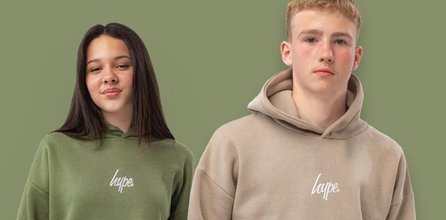 Just Hype fashion image showing a young man and woman wearing green and beige hoodies that prominantly feature the brand logo
