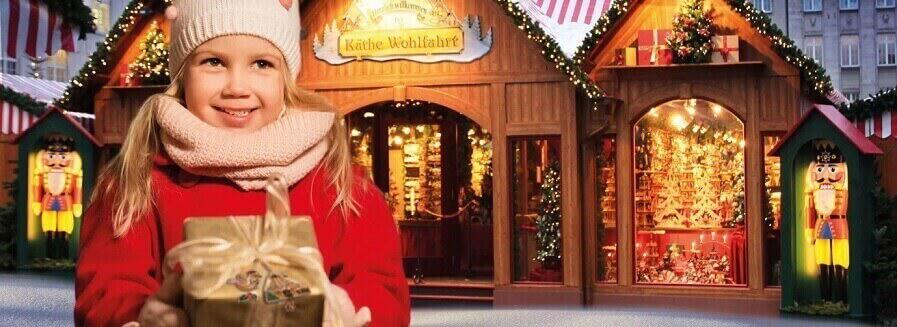 Kleines Mädchen präsentiert ein Geschenk vor dem Weihnachtsmarktstand als Banner der Käthe Wohlfahrt Gutscheinseite