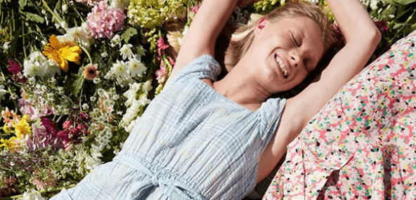 marks & spencer spring and summer savings - Image shows a woman in a light blue dress with her arms above her head enjoying the sunshine, while lying on flowers