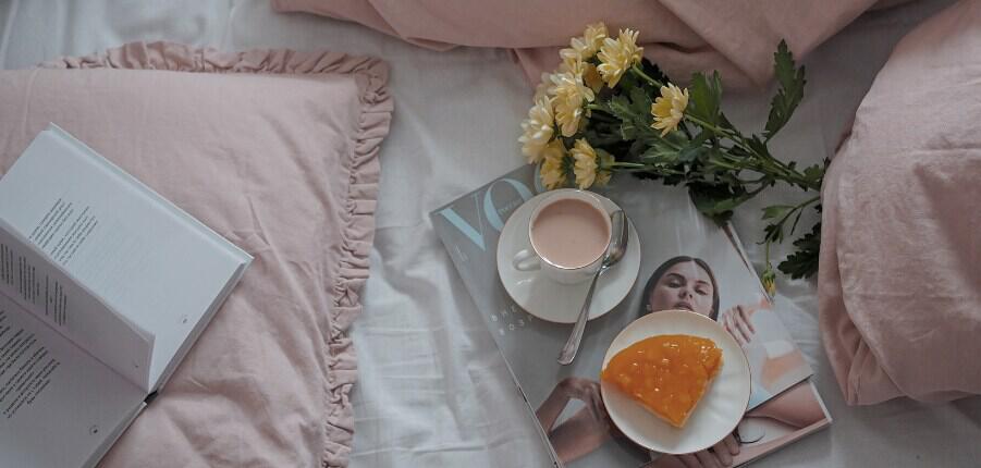 New Year sel-care image showing a magazine, flowers, cake, cup of tea and open book surrounded by pale pink cushions, 901x430px