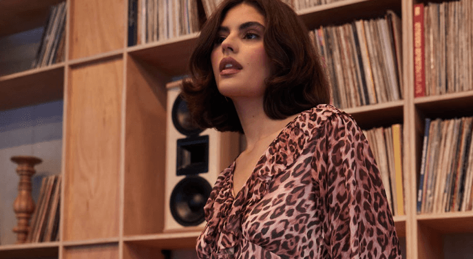 Oasis new season image showing a female model in a record store wearing a pink tinged leopard print dress