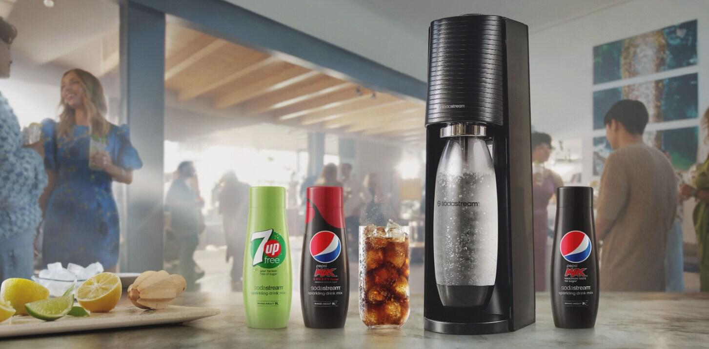 Sodastream how to save money banner - A Sodastream machine and falvour bottles on a wooden table with a social gathering in the background