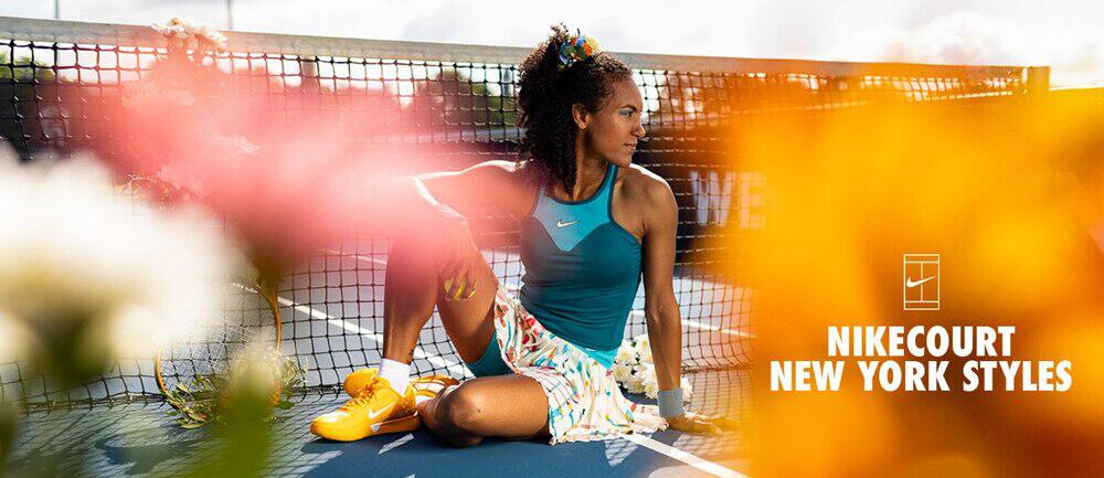 Frau sitzt am Netz auf dem Tennisplatz und trägt Top und Tennisrock aus der Nike New York Kollektion, die bei Tennis Point erhältlich ist