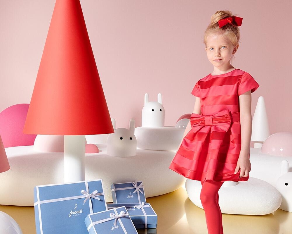 Petite fille blonde habillée dans une tenue de Noël rouge Jacadi devant des paquets cadeau