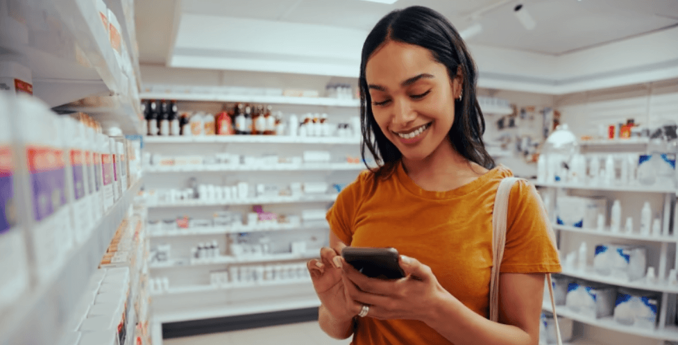 The Independent Pharmacy discount guide banner - A smiling woman looking at her mobile in front of pharmacy sheves stocked with products including pill bottles