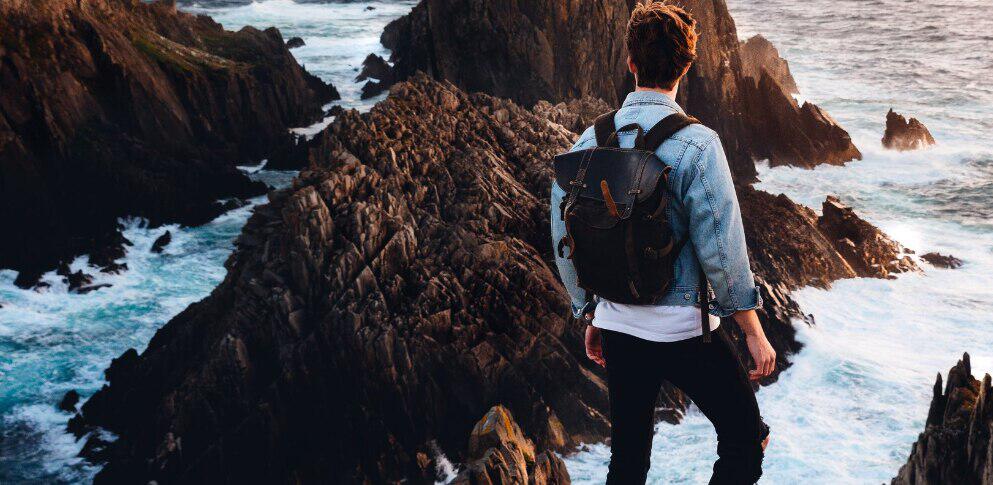 Trail Outdoor Leisure ways to save banner - A man in denim jacket and brown backpack overlooking a rugged coastal view