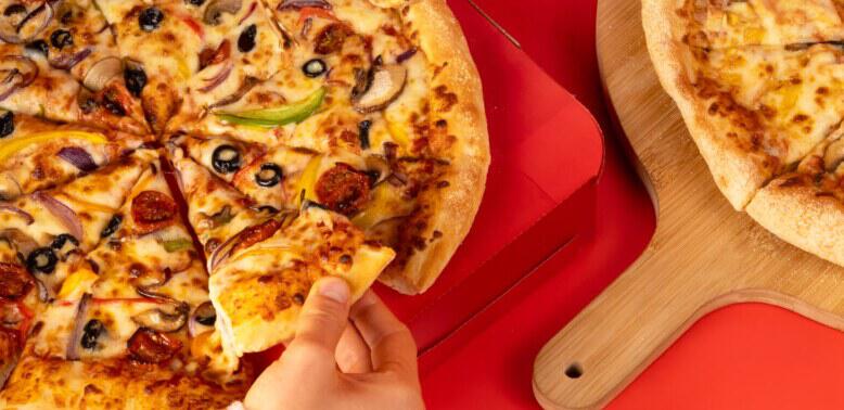 Picture of a hand taking a slice of a Plant Based Veg Supreme Pizza